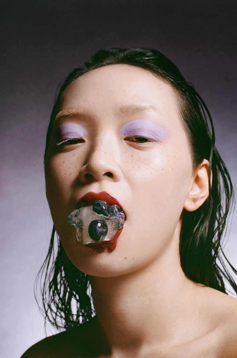 Close-up portrait of an Asian female model with dramatic makeup and blueberries in her mouth, set against a gradient grey background.