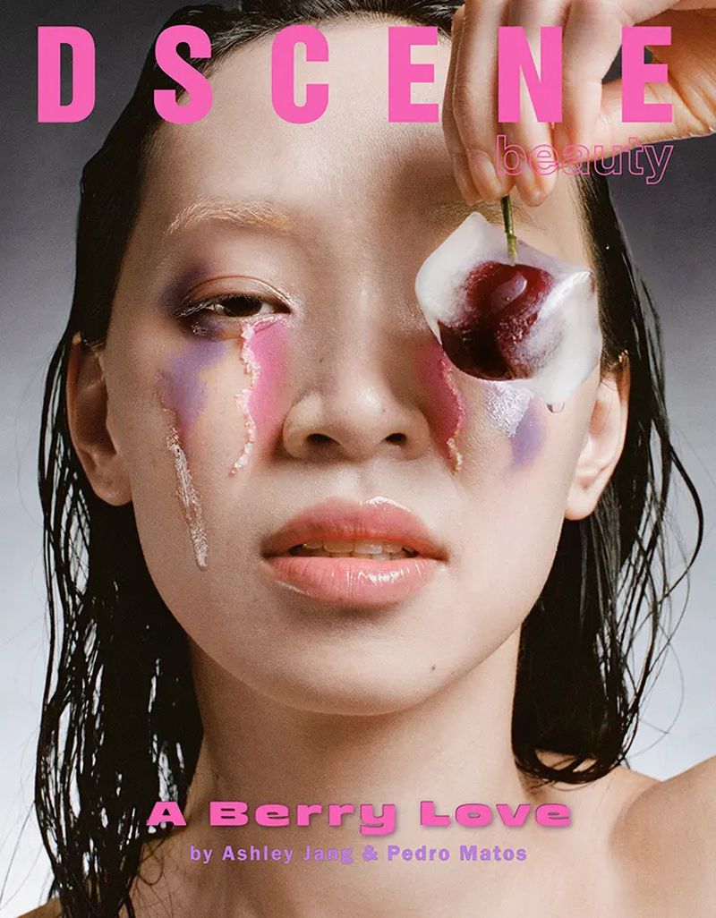 Magazine cover featuring a woman with creative pink and purple makeup, holding a cherry with ice on her eye in a close-up shot.