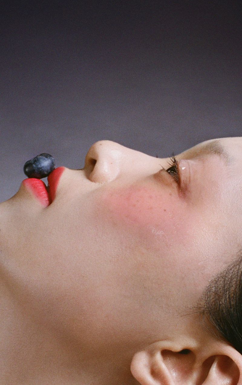 Close-up of a woman with red lipstick lying on her back with a blueberry on her lip against a grey background. Makeup and skincare concept.