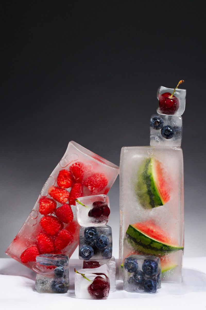 Still life of ice cubes encasing fresh fruit including watermelon, raspberries, cherries, and blueberries on a white surface.