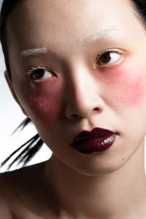 Portrait of a woman with dramatic makeup: bold lipstick, pink blush, and white eyebrows in a close-up studio setting.