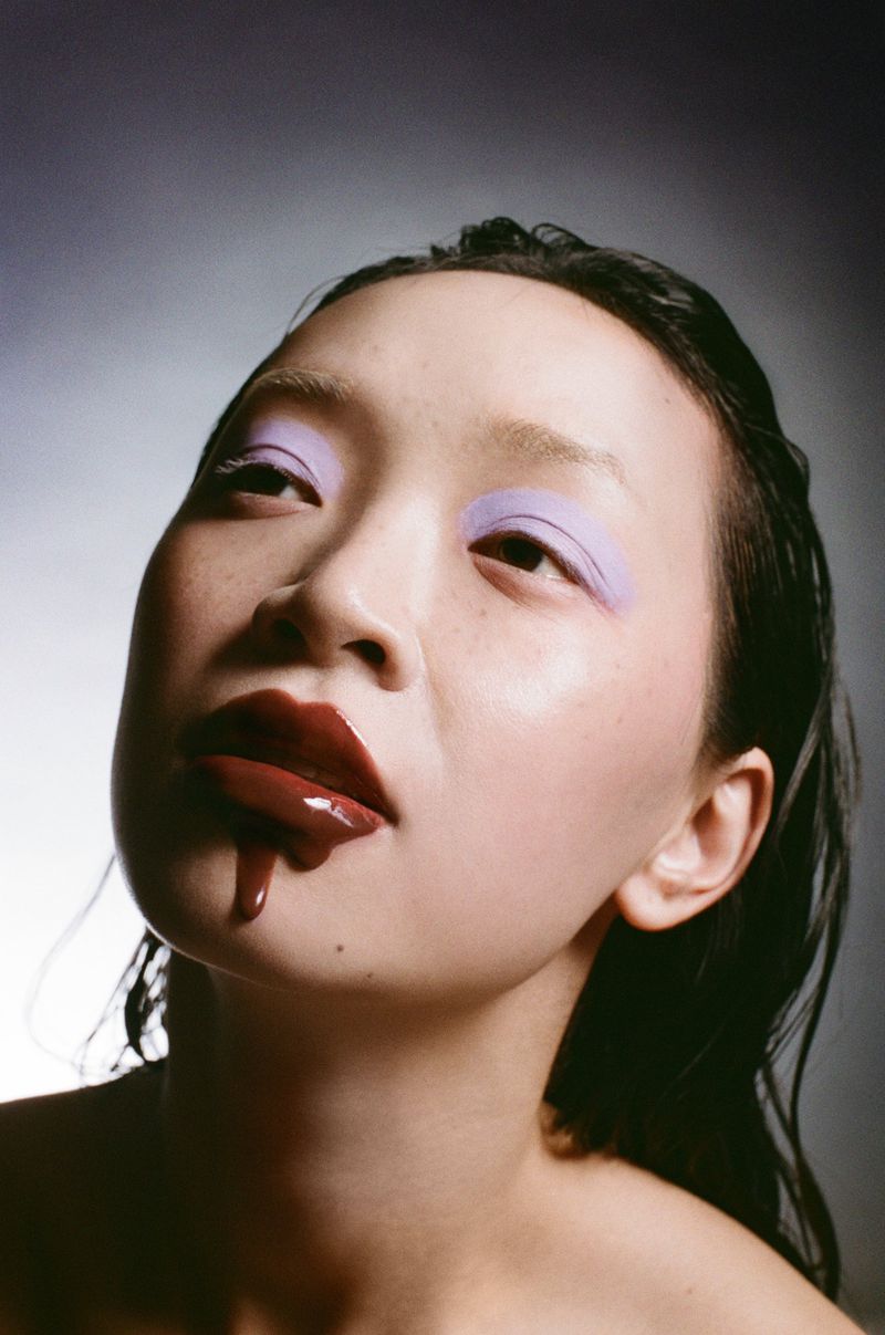 Close-up portrait of a young Asian woman with dramatic purple eyeshadow and dripping dark red lip gloss, set against a dark backdrop.