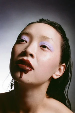 Close-up portrait of a young Asian woman with dramatic purple eyeshadow and dripping dark red lip gloss, set against a dark backdrop.