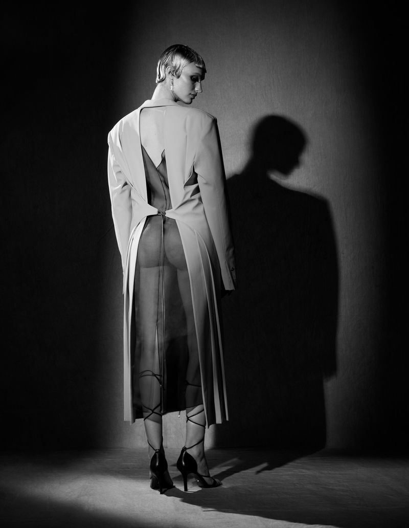Black and white fashion shot of a person with short blonde hair in a light coloured coat, sheer dress, and black heels, against a dark wall.