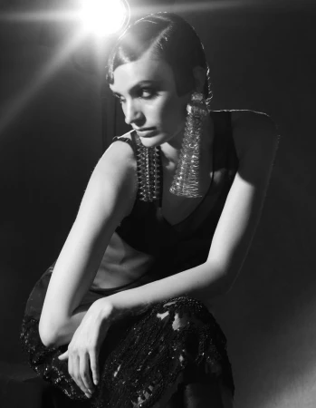 Monochrome fashion shot of a woman with vintage hairstyle wearing statement earrings and a studded top in a dark studio setting.