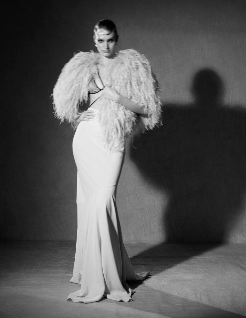 Black-and-white portrait of a fashion model in a white gown with feathered shawl, striking a pose with a serious expression in a studio setting.