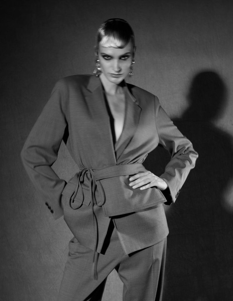 Black and white portrait of a woman in a belted suit, posing with a hand on her hip against a studio backdrop with soft lighting.