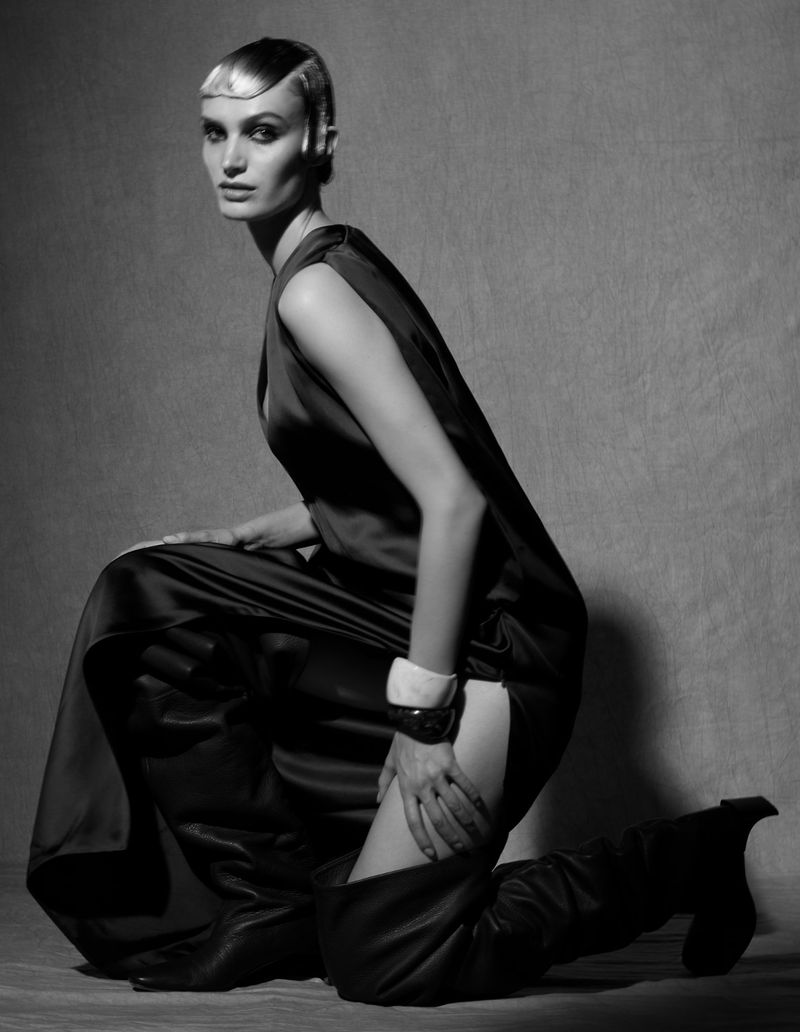 Black and white portrait of a woman in a dark dress kneeling with thigh-high boots, jewelry, and retro-styled blonde hair.