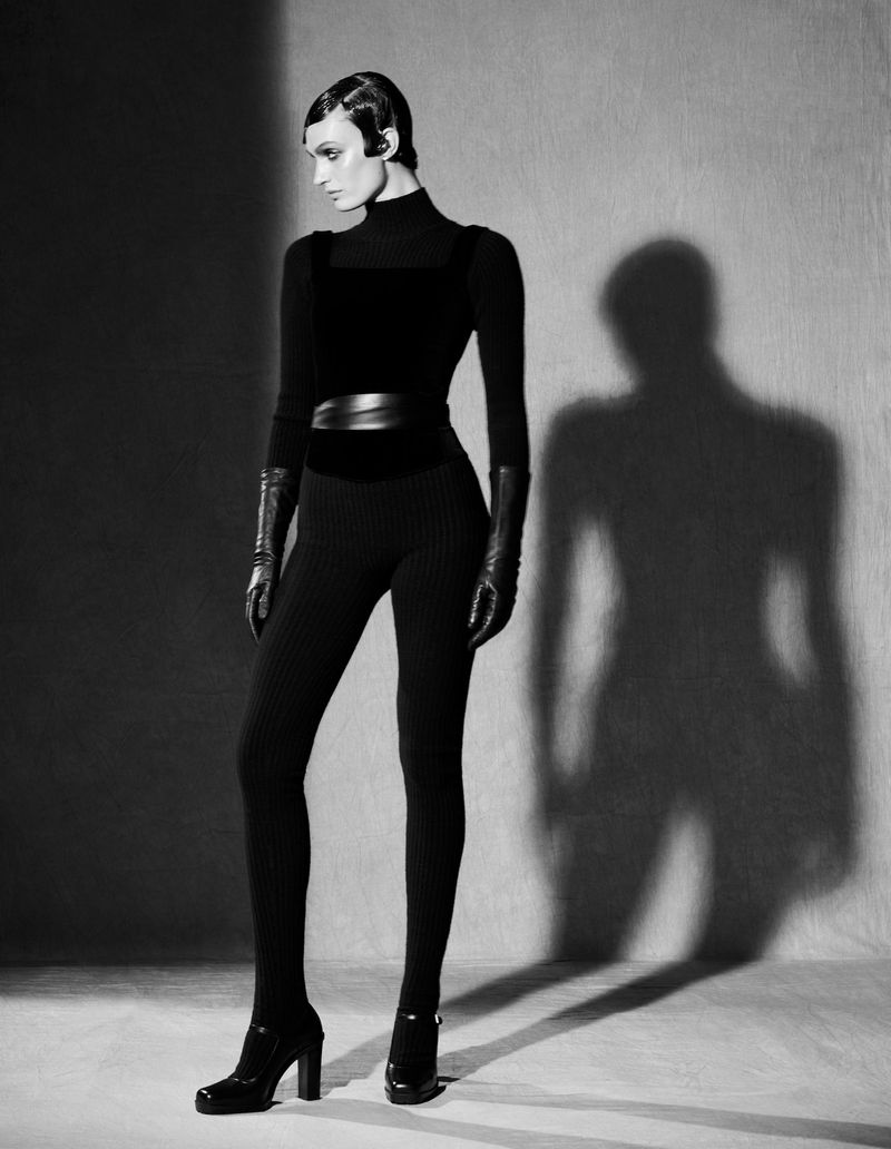 Black and white studio shot of a woman in a black jumpsuit and high heels, posing in front of a grey backdrop with her shadow.