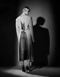 Model with short hair wearing a cutout blazer and sheer dress standing against a dark wall with a prominent shadow.