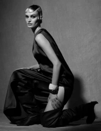 Model in dark sleeveless dress and tall boots kneeling against a textured grey studio background