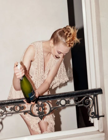 Woman in lace dress pops champagne near a railing. The scene features a creative hairstyle and a celebratory mood.