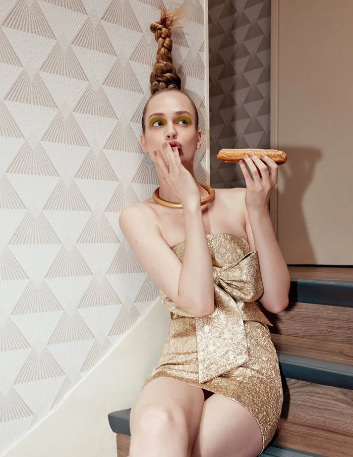 Model in a gold dress holding an eclair on stairs against patterned wallpaper for Numero Russia editorial.