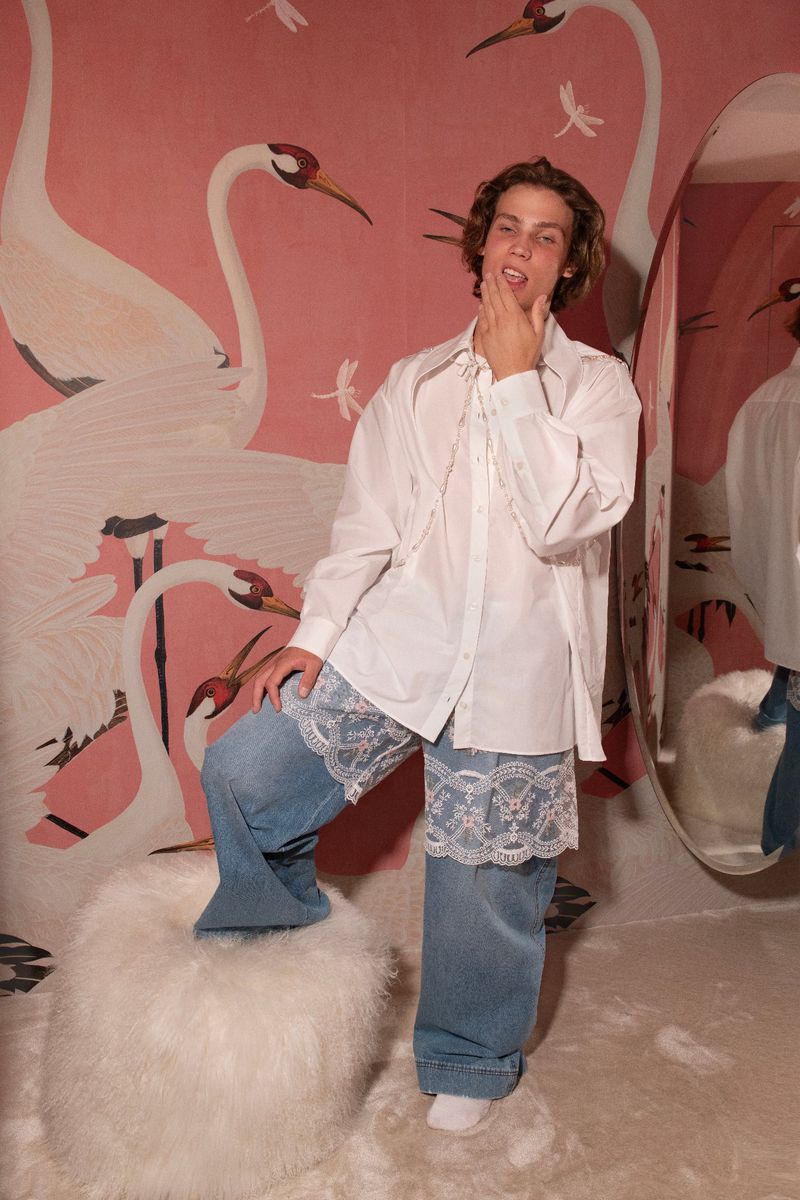 Young adult posing indoors with light-pink wall displaying large white cranes and dragonfly art, wearing white button-down shirt and jeans.