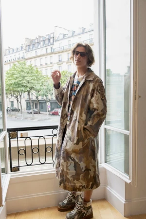 A person in a camouflage coat and sunglasses stands by a window in a building, with other buildings visible in the background.