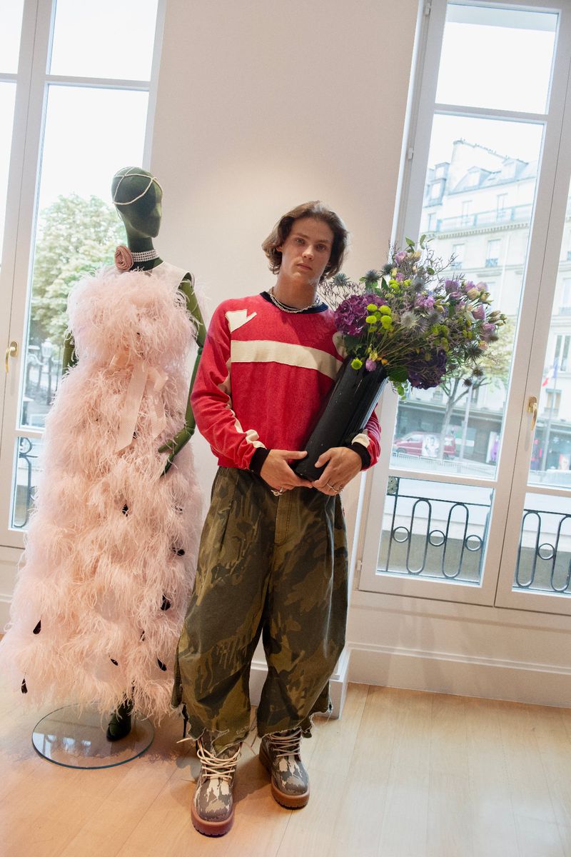 Young adult man in patterned pants holds flower arrangement next to fashion mannequin in a showroom with daylight windows.