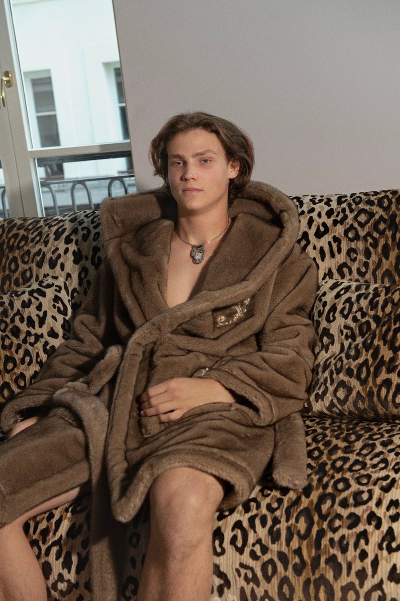 A young adult male with auburn hair sits on a leopard-print sofa wearing a brown faux fur robe with a hooded collar indoors.