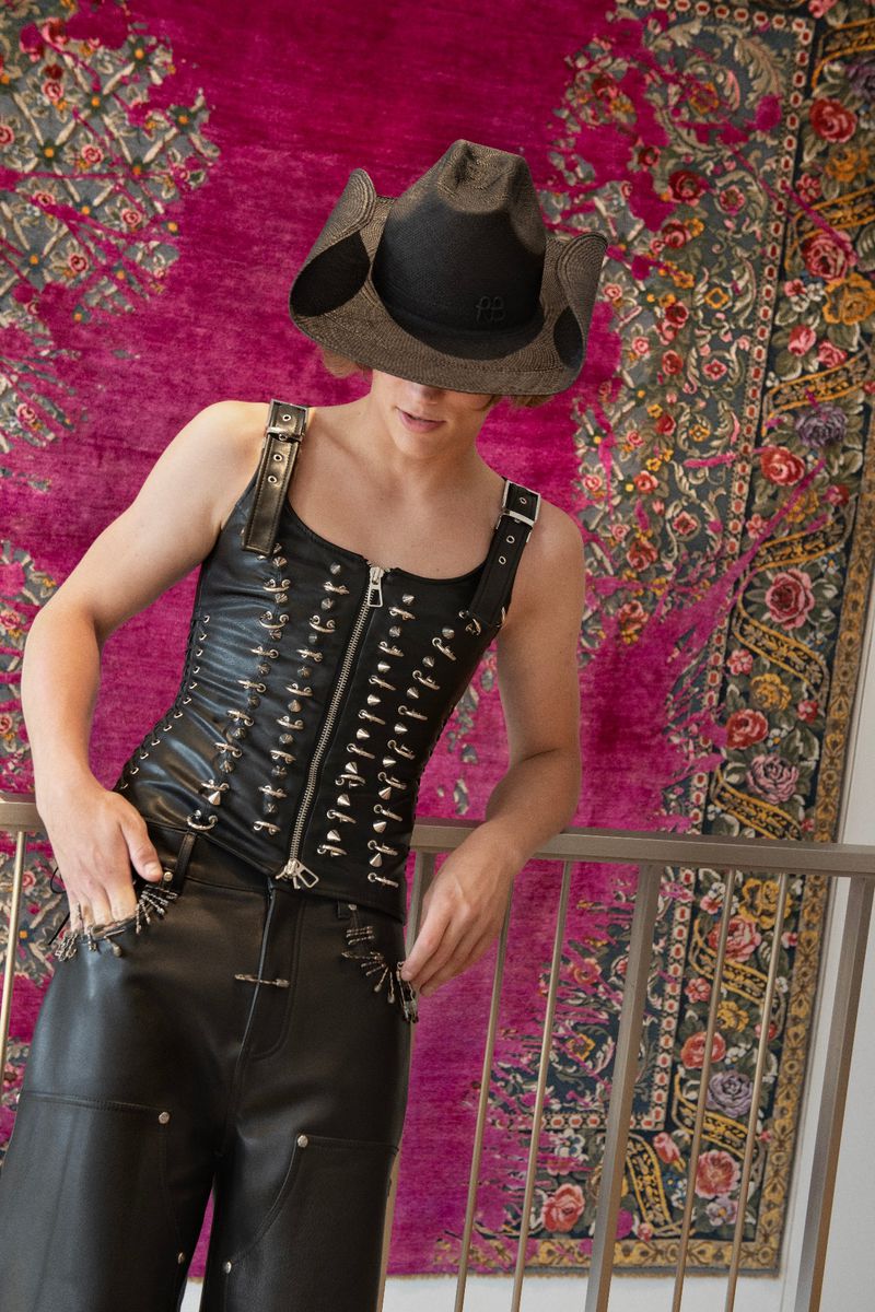 Person in a black cowboy hat and leather corset leans against a railing with a colorful rug visible in the background in a medium shot.
