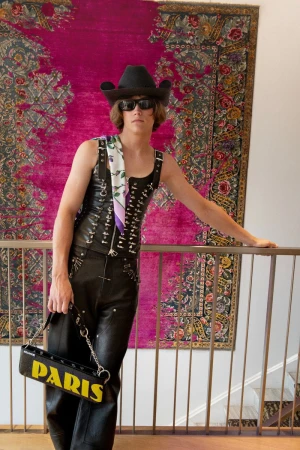 An adult male poses in a leather outfit with a black cowboy hat, handbag, and sunglasses against a colorful rug indoors.