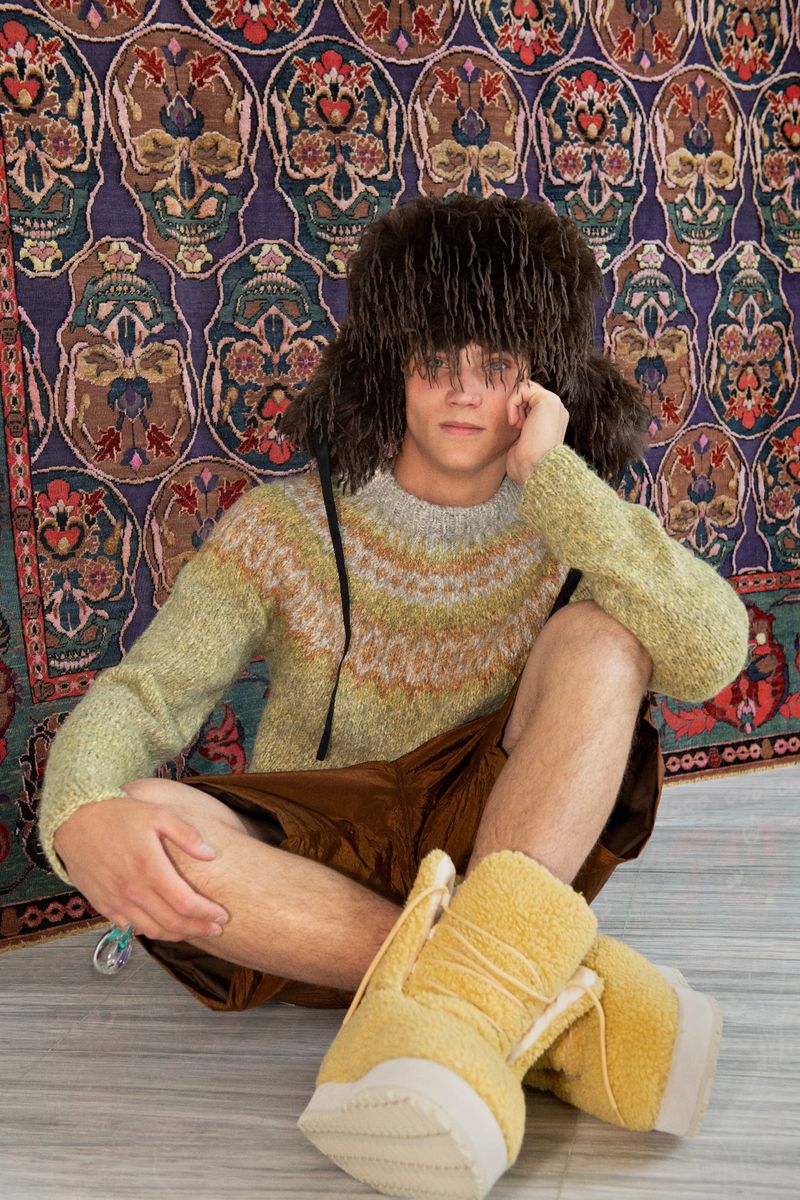 Young adult in fuzzy hat, patterned sweater, and mustard boots poses indoors against a patterned skull tapestry backdrop.