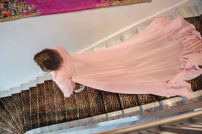 Overhead shot of a woman in a pink dress walking down carpeted stairs, dress flowing, creating a soft, elegant visual narrative.