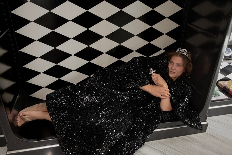 Person lies in reflective interior wearing sequin jacket and tiara, posed on black and white checkerboard floor, indoor studio setting.