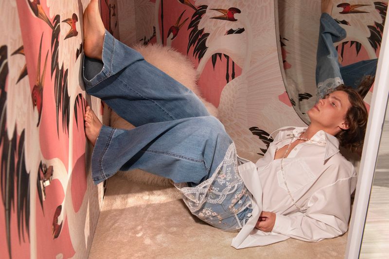 A fashionable young woman lies on an upholstered surface in blue jeans and white shirt with a striking bird-themed wallpaper background.