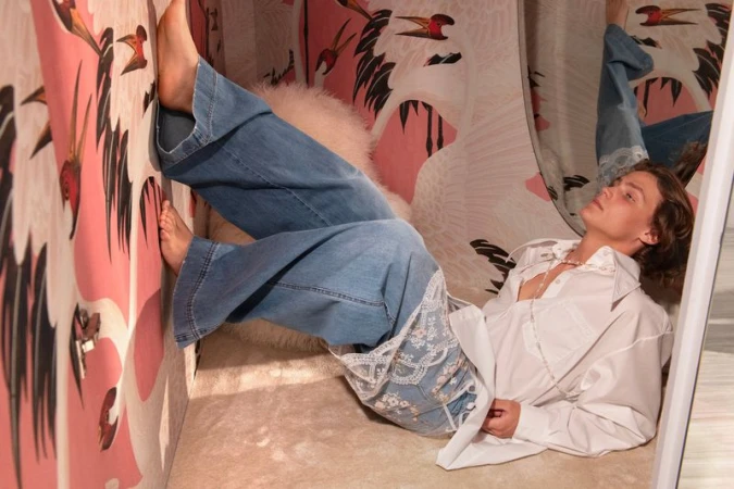 A fashionable young woman lies on an upholstered surface in blue jeans and white shirt with a striking bird-themed wallpaper background.