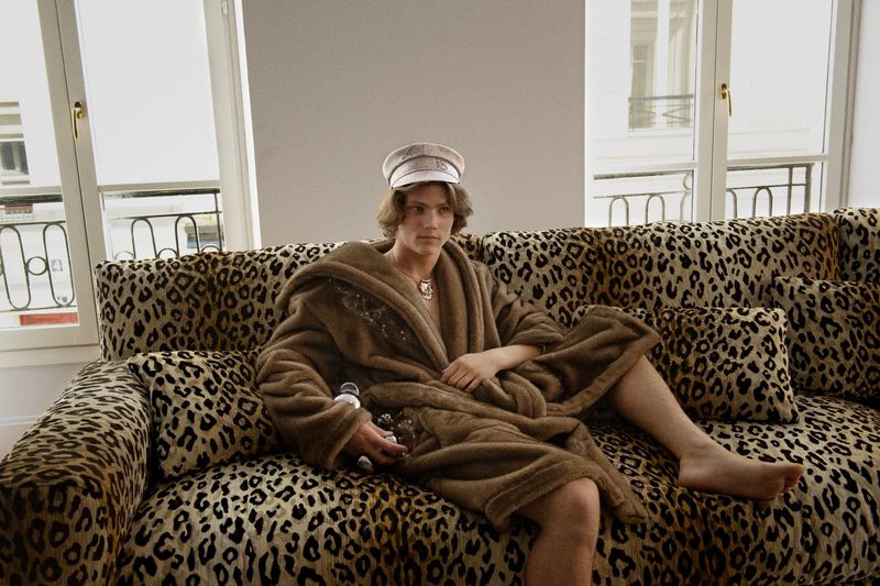 Man in robe sits on an animal print couch indoors. Natural light from windows illuminates the setting, creating a relaxed, casual scene.