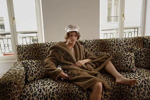 Young man in a brown plush robe and patterned cap seated on a leopard print couch before bright windows.