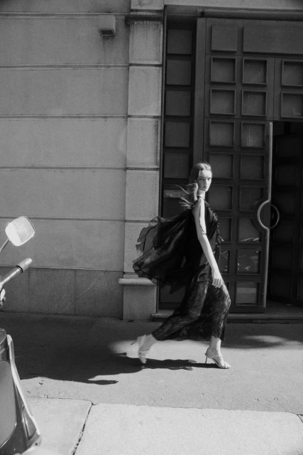 Monochrome fashion shot of a woman in a dress and heels walking in front of a building with a moped on the sidewalk.