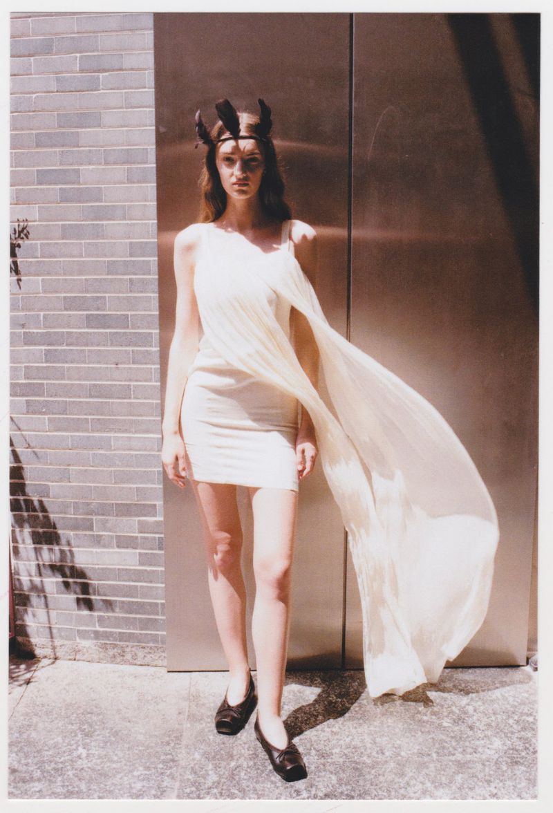 A slender woman poses in a light-colored dress and dark flats against a brick wall and metal door, casting a shadow in an outdoor setting.