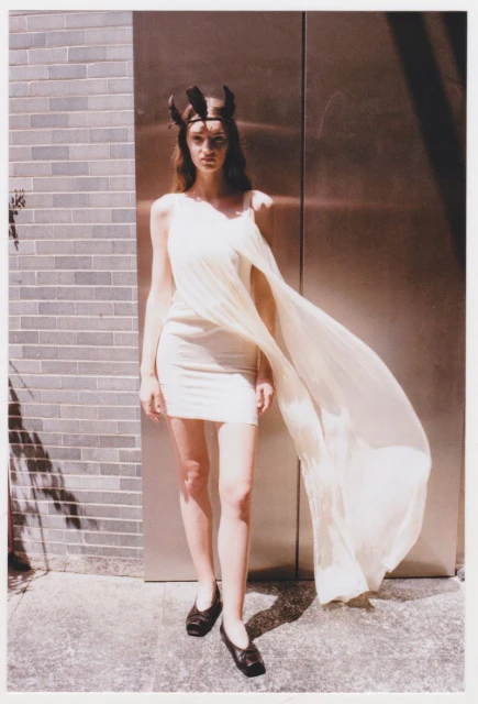A slender woman poses in a light-colored dress and dark flats against a brick wall and metal door, casting a shadow in an outdoor setting.