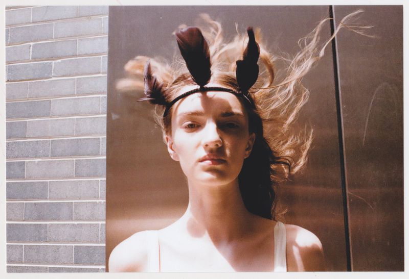 Portrait of a female model with a feather headpiece, striking a pose with neutral expression, indoor lighting, and a gray brick wall.