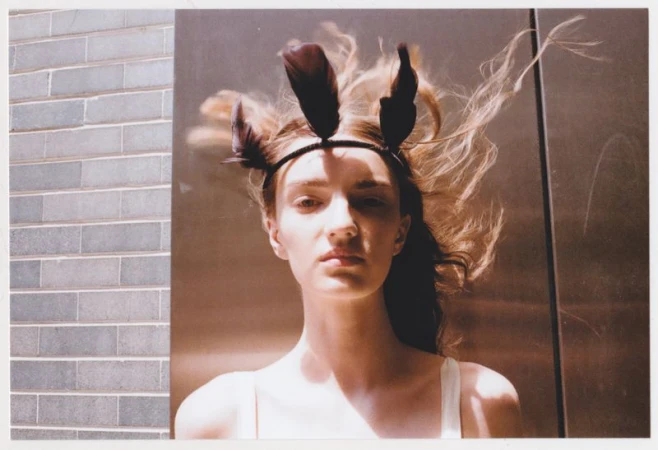 Portrait of a female model with a feather headpiece, striking a pose with neutral expression, indoor lighting, and a gray brick wall.