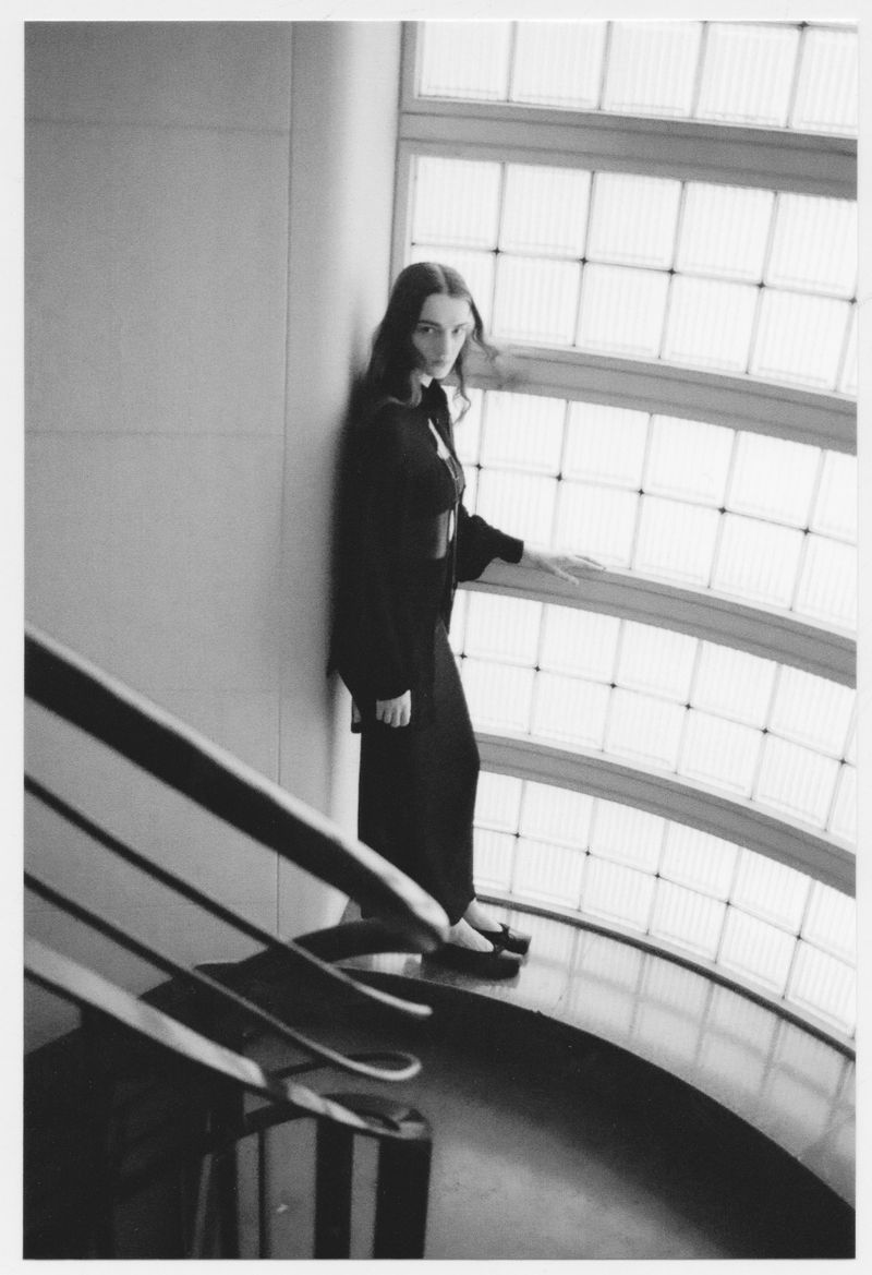 Monochrome portrait of a woman standing indoors by a window with patterned glass, wearing a dark jacket and pants, captured in soft light.