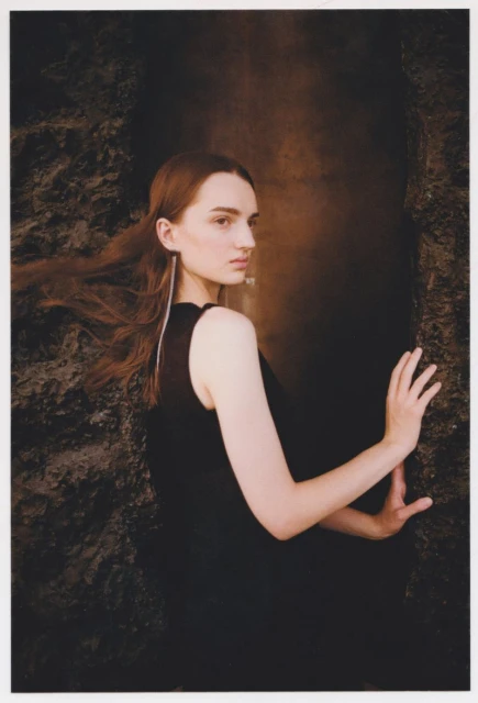 A young woman poses in a dark sleeveless top, set against a textured stone wall and a dark backdrop with subtle reddish-brown lighting.
