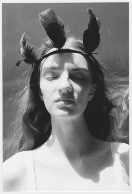 Monochrome portrait of a fair-skinned young woman with closed eyes, wearing a feathered headband, against a blurred background.