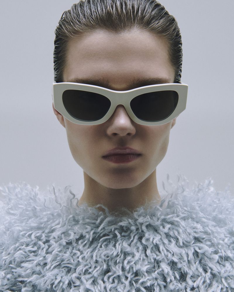 Close-up shot of a model wearing white sunglasses and a light blue fuzzy top against a neutral background, demonstrating a fashion-forward aesthetic.