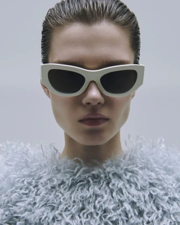 Close-up shot of a model wearing white sunglasses and a light blue fuzzy top against a neutral background, demonstrating a fashion-forward aesthetic.
