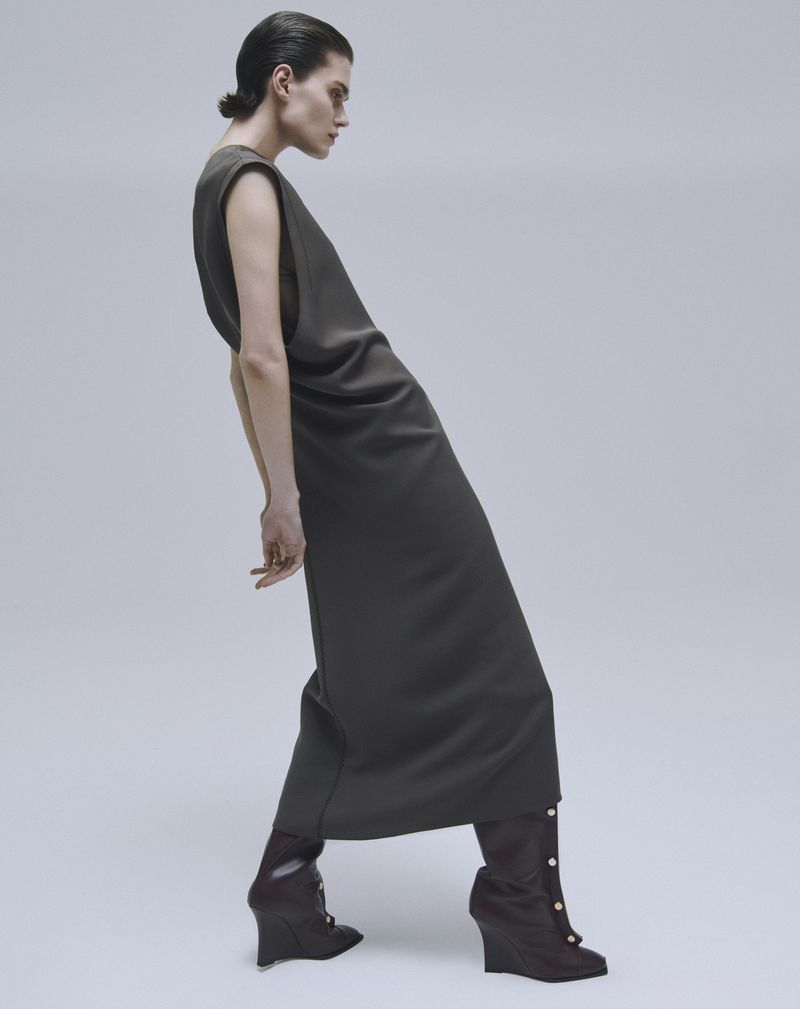 Fashionable woman in a sleeveless olive-green dress with pointed-toe wedge ankle boots poses in a studio with a light-grey background.