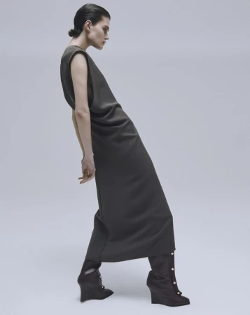 Fashionable woman in a sleeveless olive-green dress with pointed-toe wedge ankle boots poses in a studio with a light-grey background.