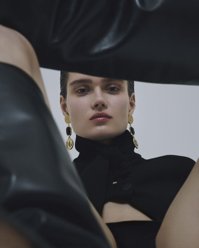Close-up shot of a fashion model with sleek dark hair, wearing a black turtleneck dress and gold drop earrings, posing against a white background.