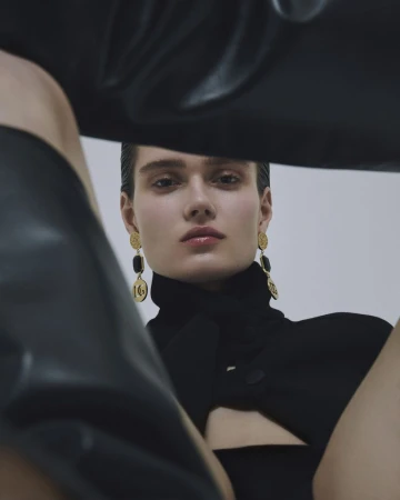 Close-up shot of a fashion model with sleek dark hair, wearing a black turtleneck dress and gold drop earrings, posing against a white background.