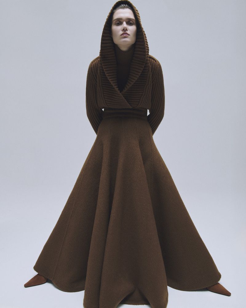 Fashion editorial shot: woman in hooded brown knit dress with long skirt, posing indoors in photo studio against a neutral background.