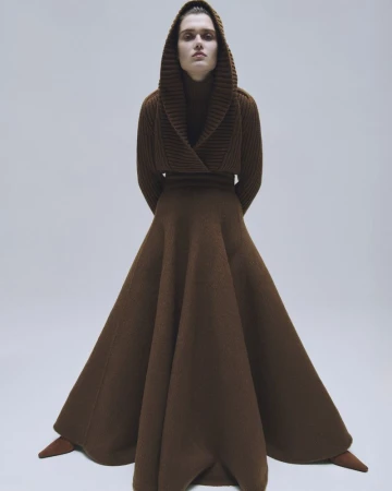 Fashion editorial shot: woman in hooded brown knit dress with long skirt, posing indoors in photo studio against a neutral background.