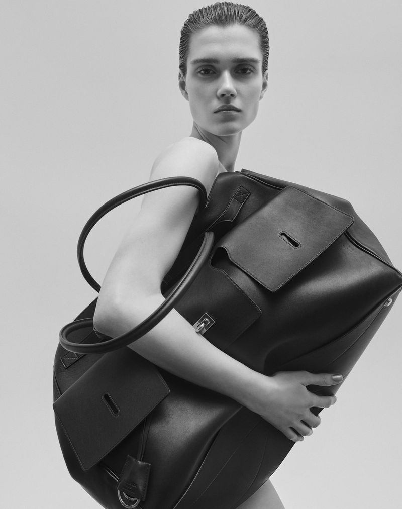 Monochrome portrait of a woman with short hair posing with a large leather duffel bag in front of a simple neutral background.