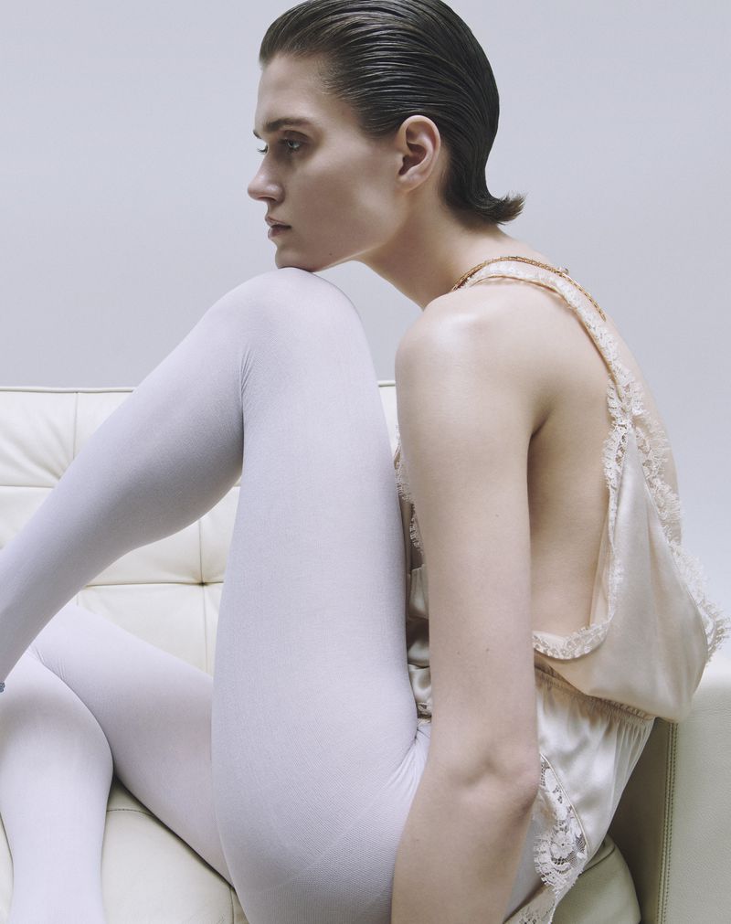 Elegant portrait of a woman in a satin top and white tights, seated gracefully against a white backdrop in a minimalist studio setting.
