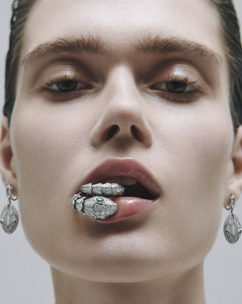 Close-up beauty shot of a model with a snake ring held in her mouth and matching snake earrings, set against a plain background.