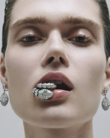 Close-up beauty shot of a model with a snake ring held in her mouth and matching snake earrings, set against a plain background.
